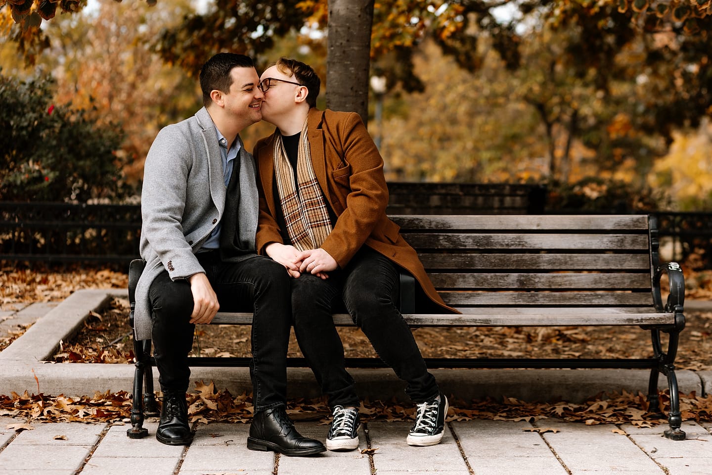 Eric and JJ kissing on a park bench surrounded by autumn leaves during their Adams Morgan engagement session in Washington, DC
