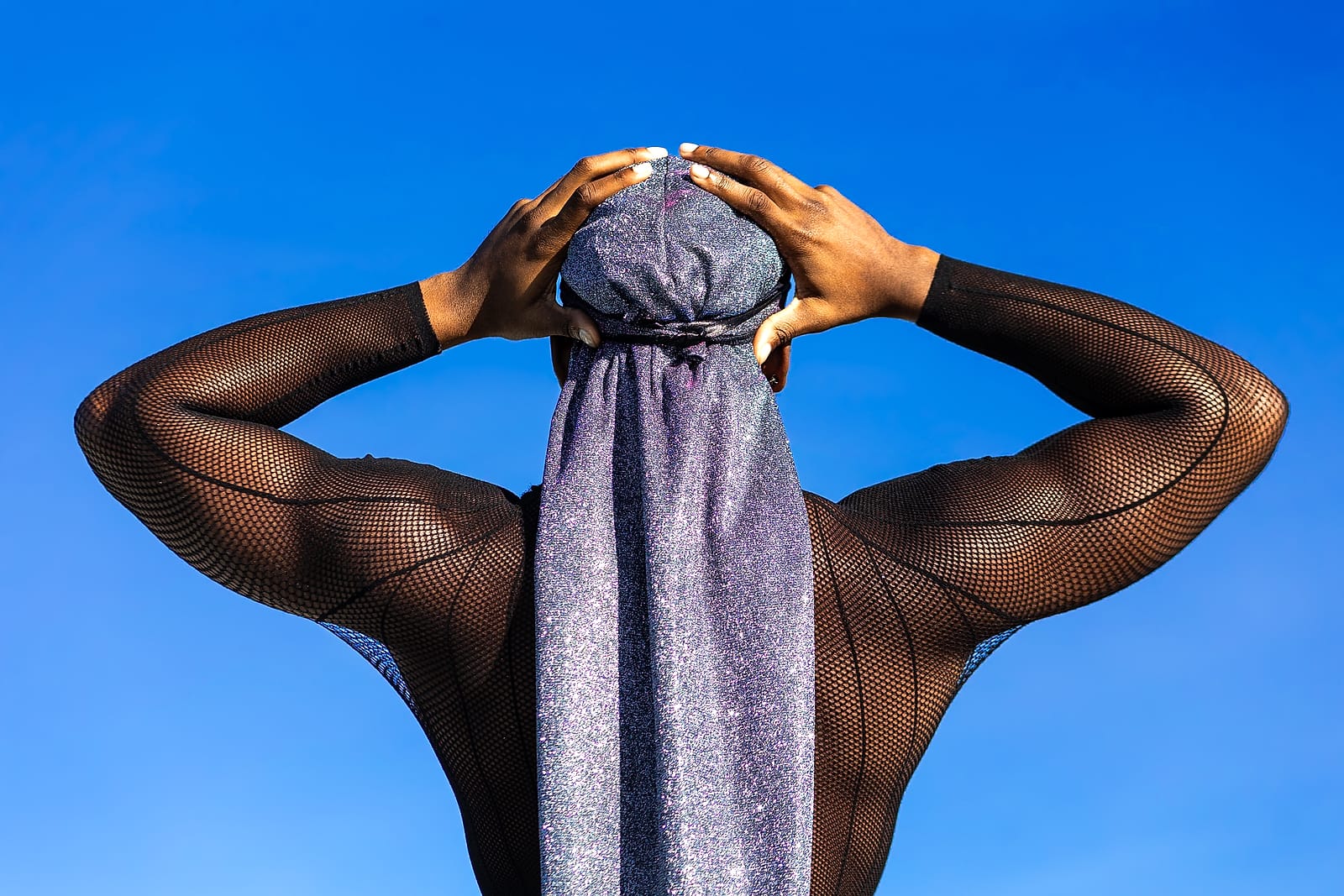 A person seen from behind against a vivid blue sky, hands framing a shimmering silver head wrap