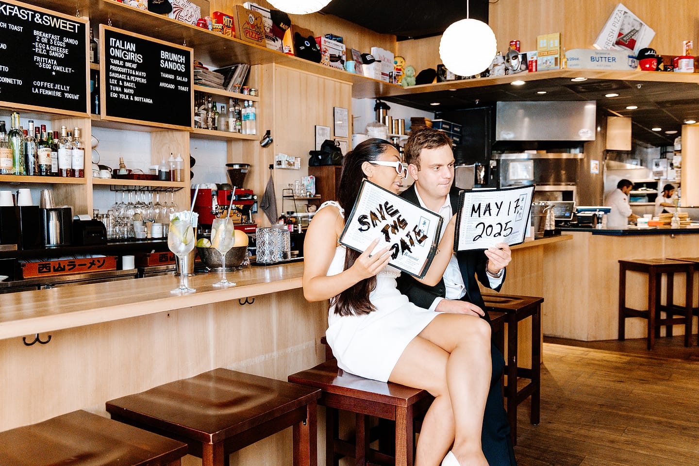 Jasmine and Aaron holding ‘Save the Date — May 17, 2025’ signs at the Tonari bar during their Chinatown DC engagement session