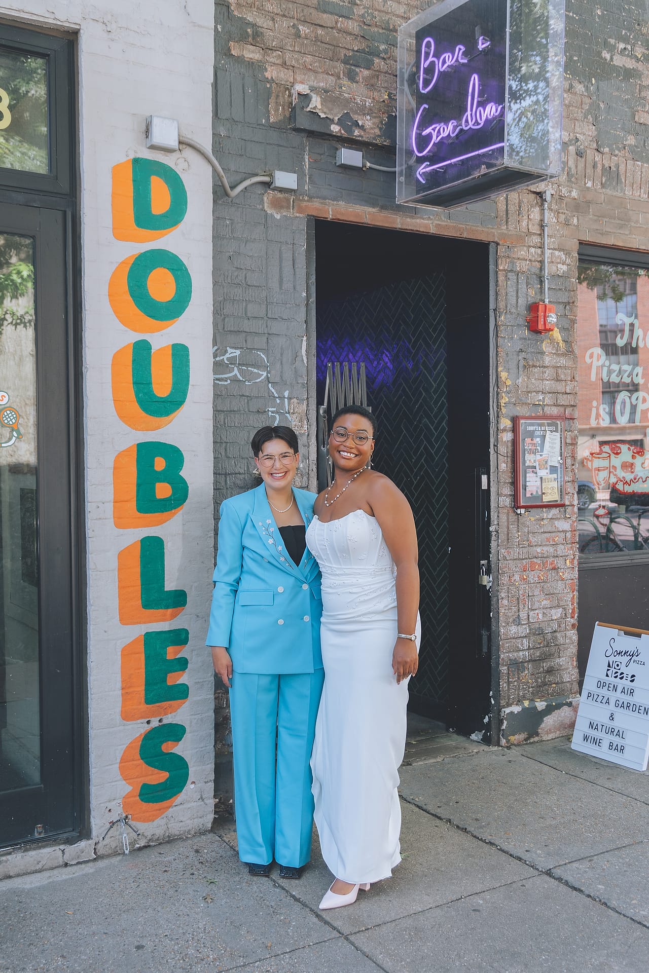 Arika and Madeline pose outside a DC bar beside a colorful Doubles mural on their wedding day