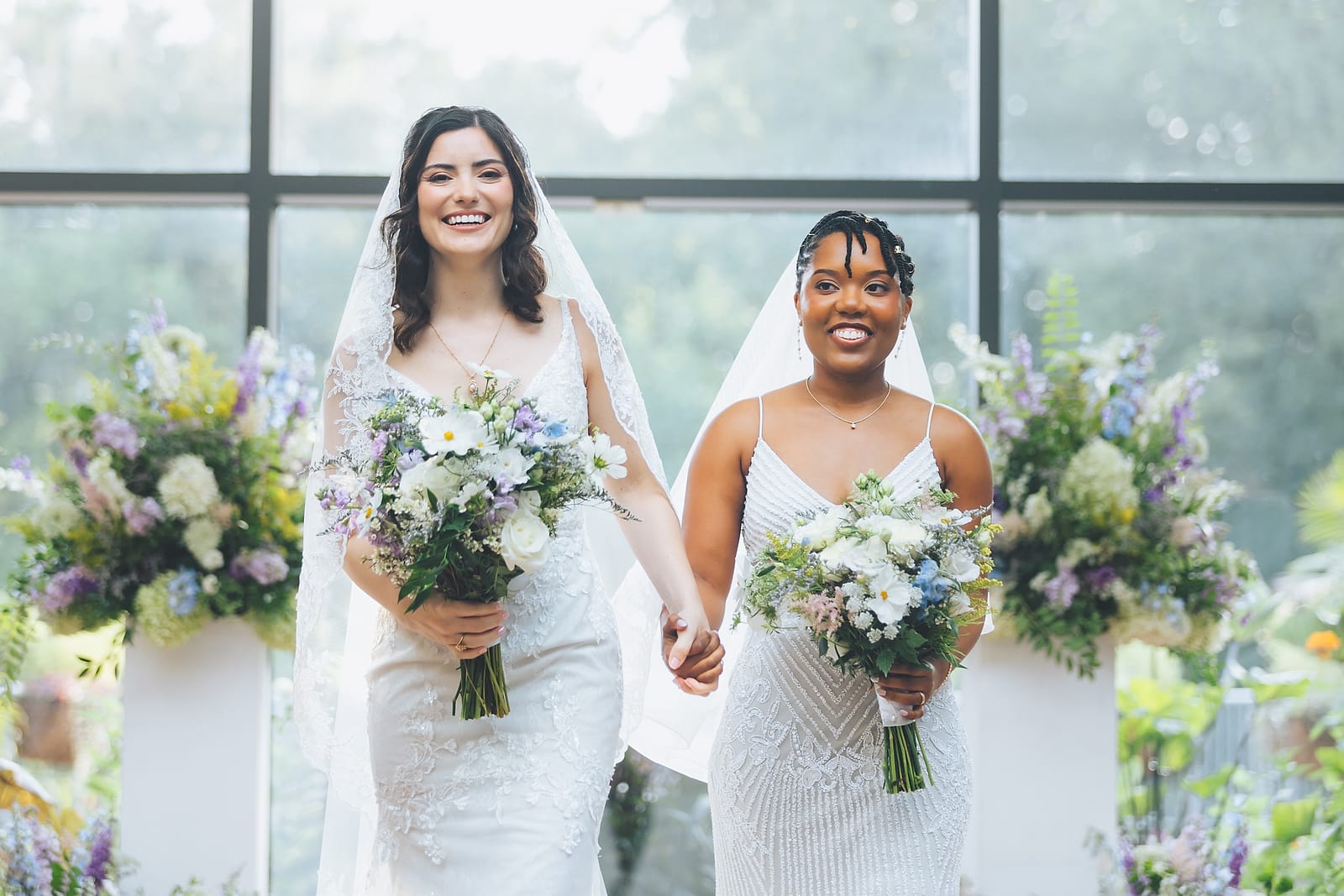 Two brides holding wildflower bouquets in a sunlit greenhouse